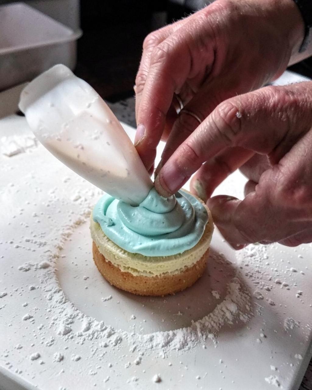 Hands piping blue buttercream onto a small cake